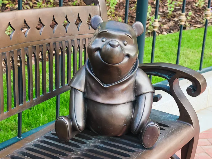 Statue en bronze de Winnie l’ourson assise sur un banc, le long d’une pelouse d’un parc Disney.