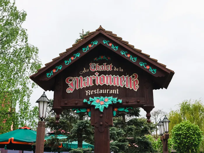 Enseigne en bois sculpté Au Chalet de la Marionnette, restaurant de Fantasyland, entourée de verdure.