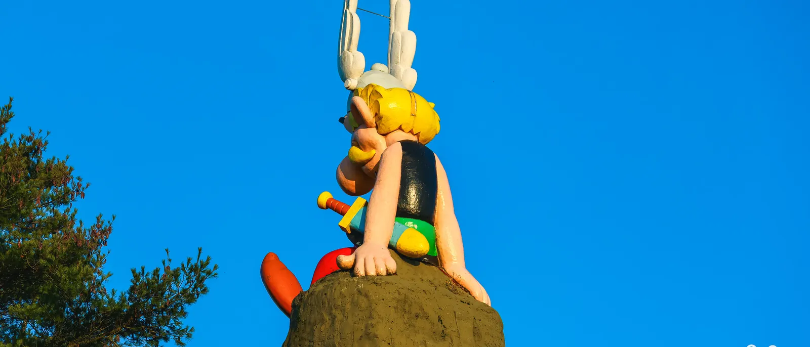 Statue d'Astérix assise au sommet d'un rocher, vue de dos en contre-plongée, casque ailé blanc sur ciel bleu