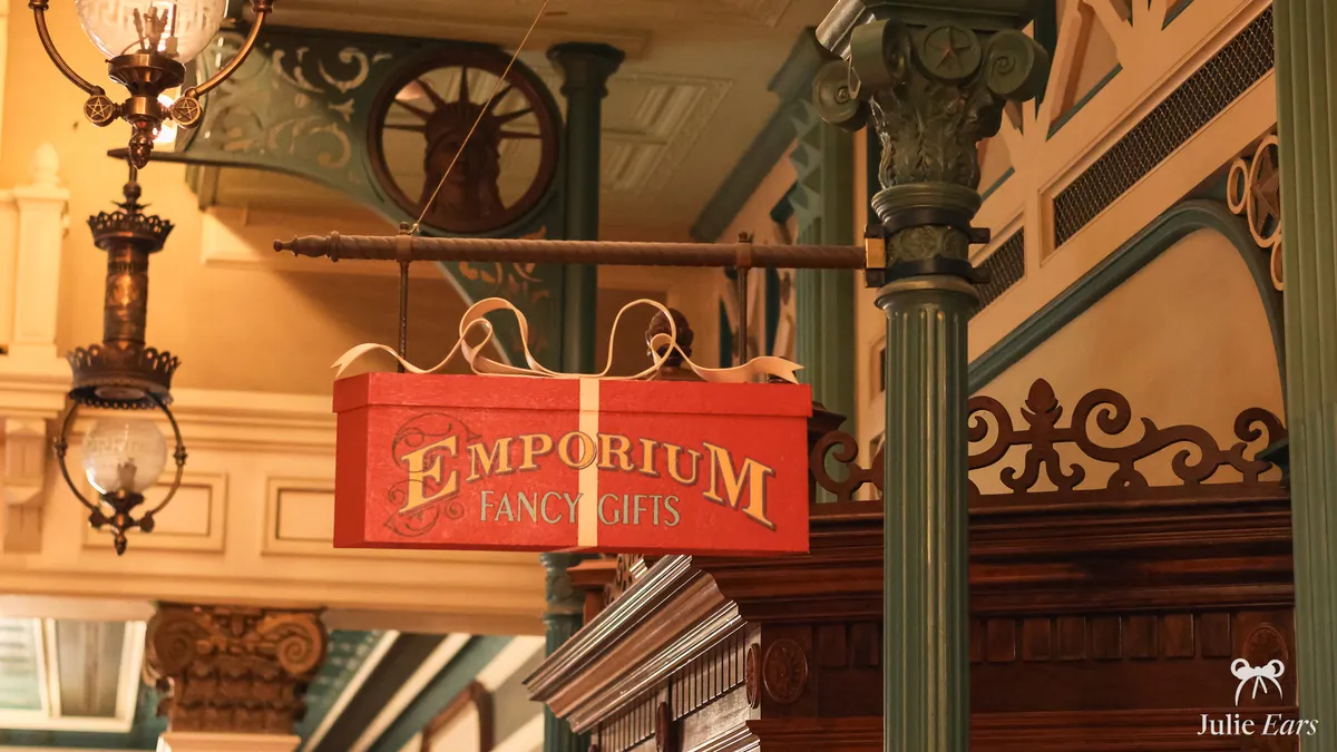 L’enseigne rouge en forme de paquet cadeau "Emporium Fancy Gifts" suspendue dans la boutique Emporium de Main Street U.S.A., entre lampes anciennes et colonnes vertes