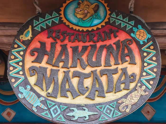 Enseigne ronde sculptée du Restaurant Hakuna Matata avec Simba et motifs africains colorés, à Adventureland.