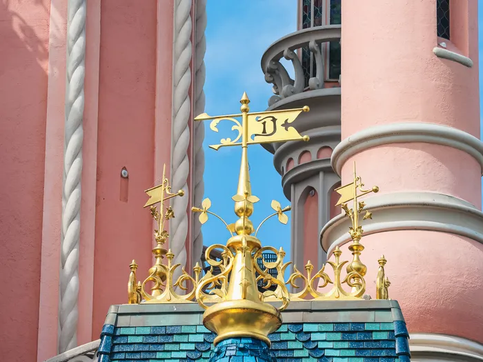 Girouette dorée marquée d’un « D » et toitures bleutées d’une tourelle rose du Château de la Belle au Bois Dormant, Disneyland Paris.