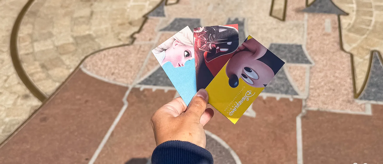 Trois billets Disneyland Paris (Elsa, Star Wars, Indestructibles) tenus en éventail au-dessus du pavage du parc.