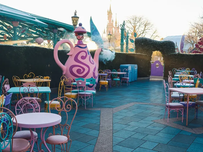 Terrasse du March Hare avec théière rose géante et chaises en fer colorées, château en fond, à Fantasyland Disneyland Paris.