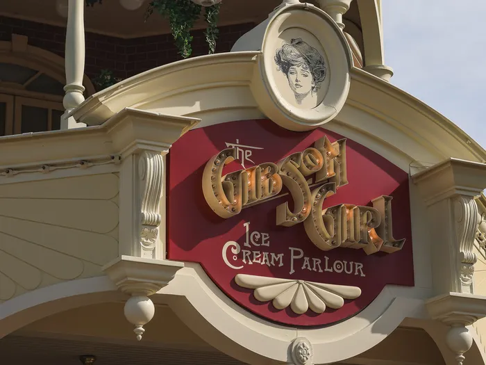 Gold-on-red sign of The Gibson Girl Ice Cream Parlour on Main Street U.S.A., topped with a drawn portrait of a young woman