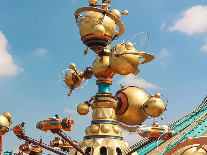 Structure dorée d’Orbitron avec ses nacelles fusées et son mât à sphères, devant le toit vert de Space Mountain à Discoveryland.