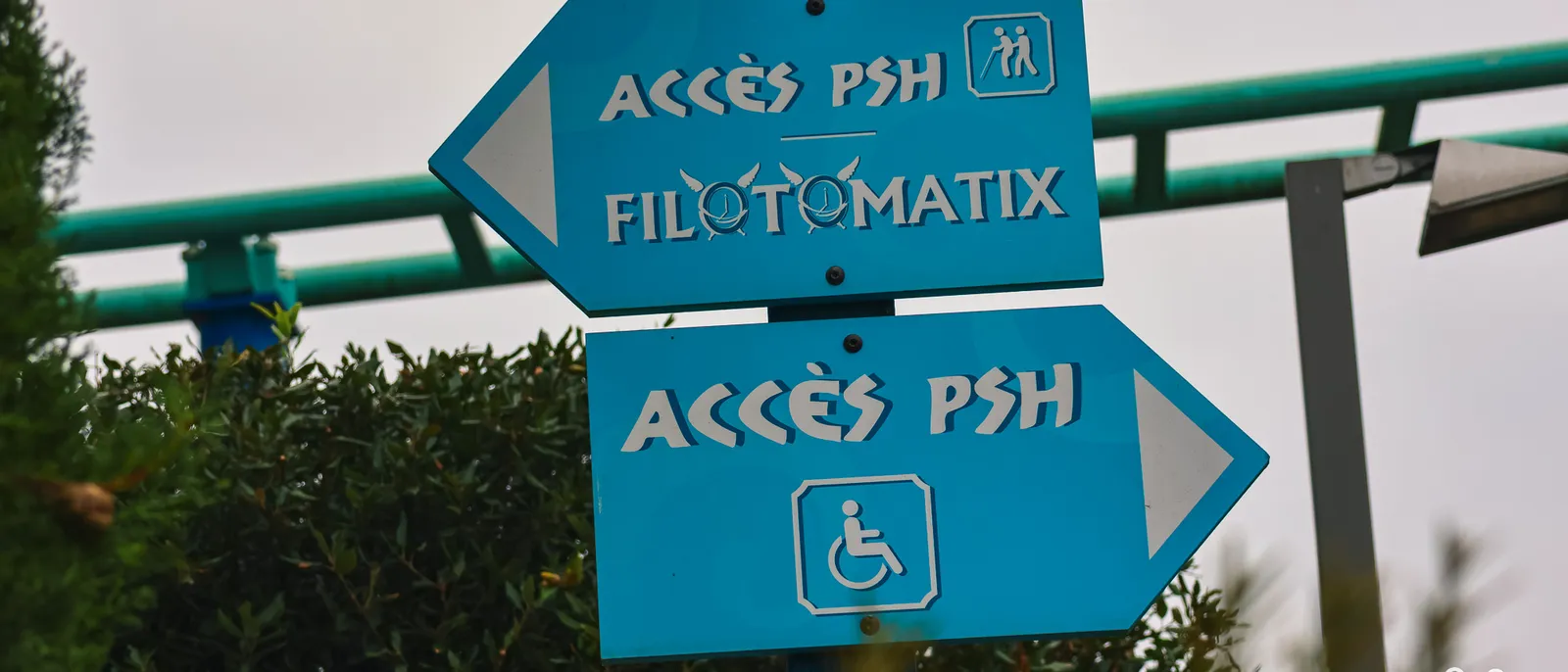 Panneaux fléchés bleus "Accès PSH" vers Filotomatix, pictogrammes fauteuil roulant, devant un rail turquoise et des haies