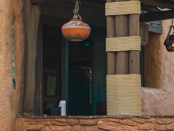 Hatch in the adobe wall of Pueblo Trading Post, under a log awning hung with a painted pot, beside a rope-bound log pillar