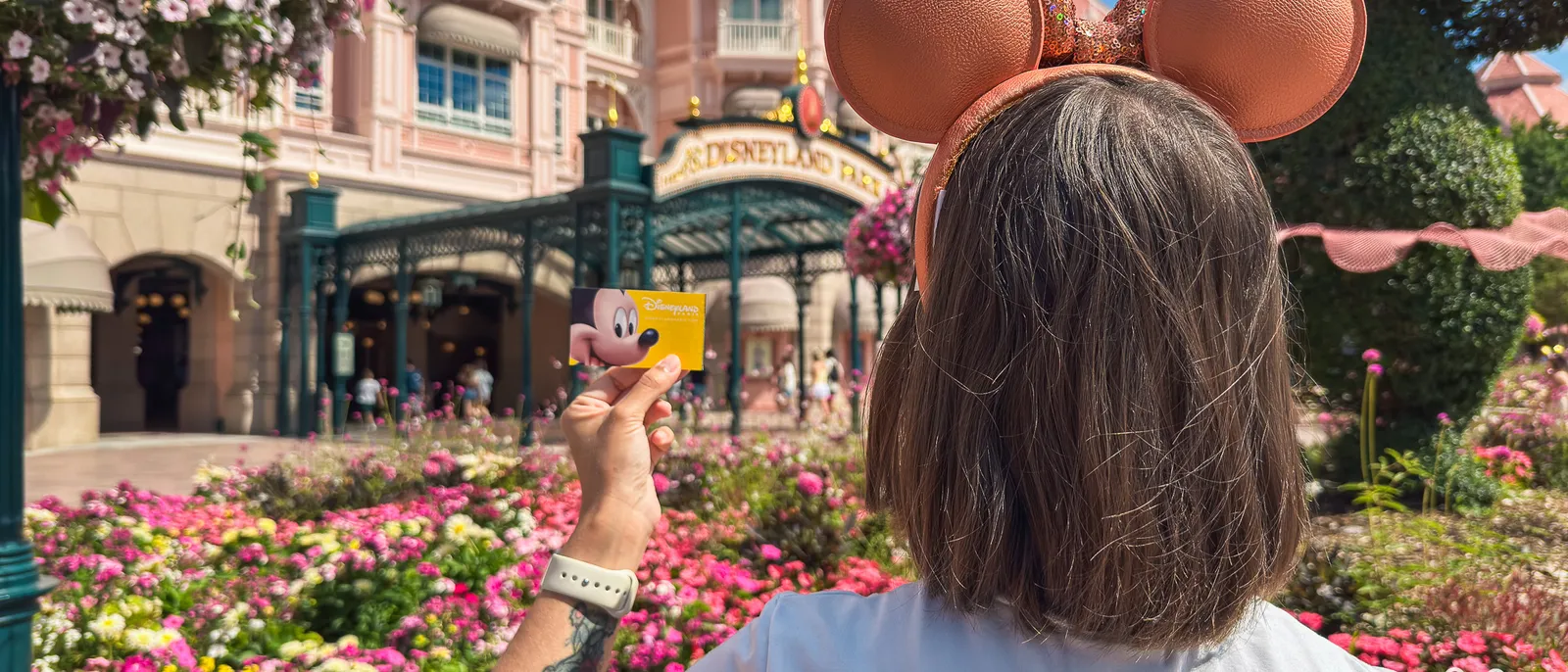 Visiteuse de dos, oreilles Minnie dorées, tenant un billet Mickey jaune devant le Disneyland Hotel et ses massifs fleuris.