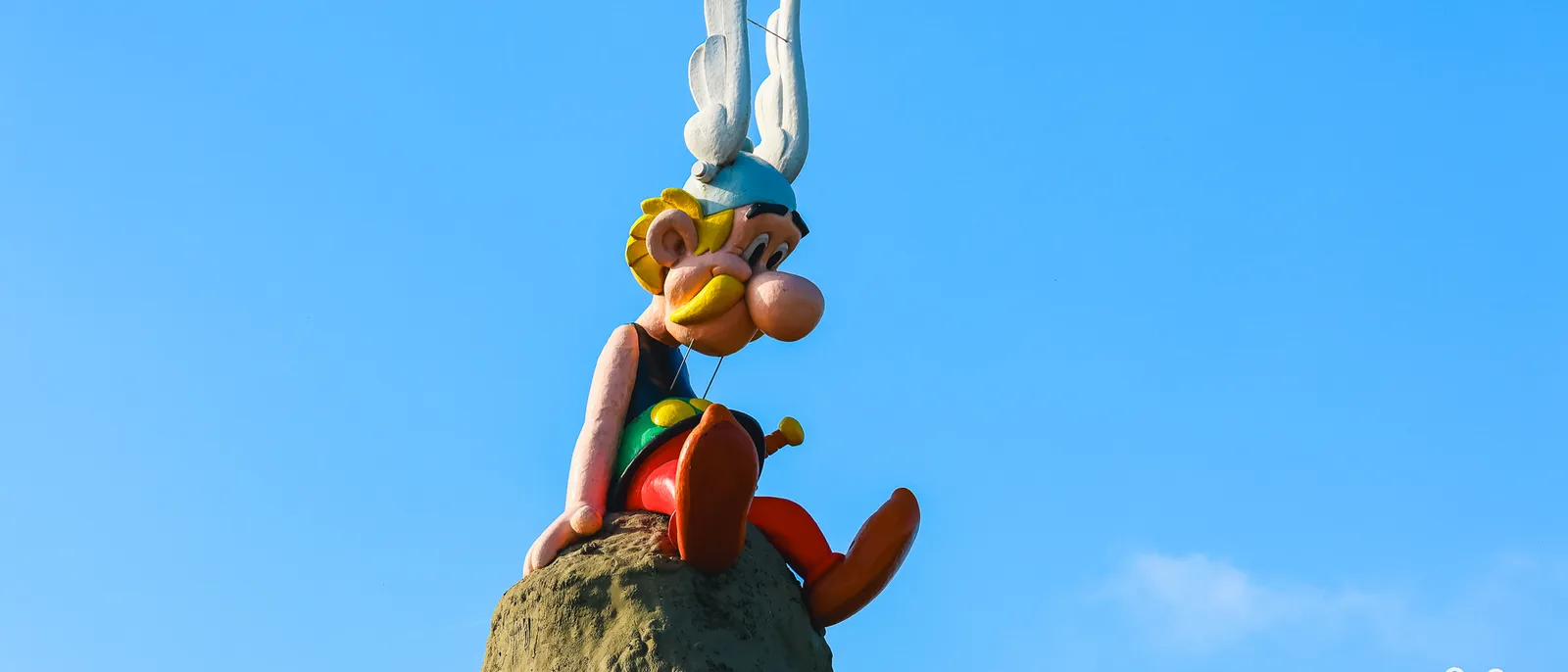 Statue d’Astérix assise sur un rocher, vue de face en contre-plongée, casque ailé et bottes rouges sur ciel bleu