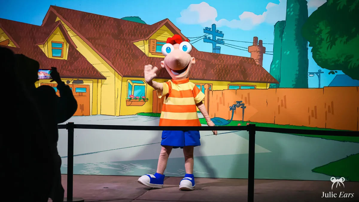 Phineas from Phineas and Ferb greeting visitors in front of a drawn backdrop of his yellow house and orange fence, at Back In Time Night 2024