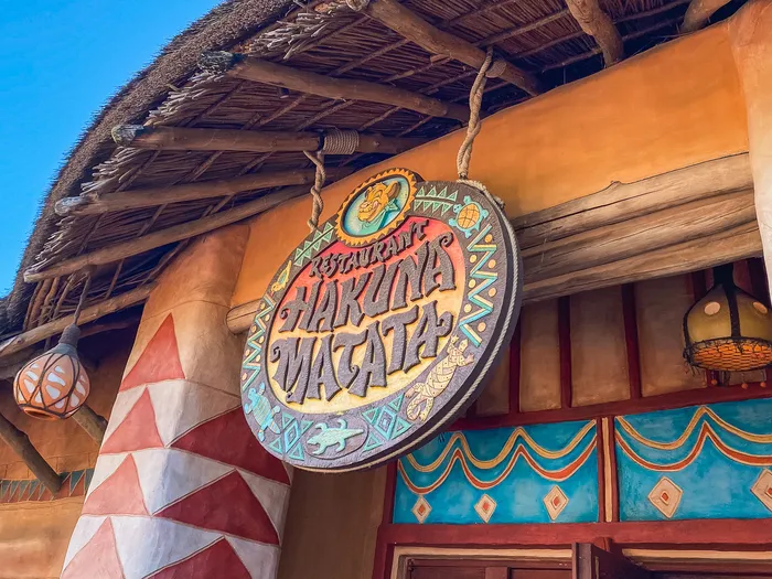 Enseigne ronde sculptée Restaurant Hakuna Matata suspendue sous un toit de chaume à Adventureland