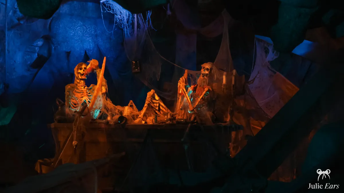 Two pirate skeletons at a table under cobwebs in the Pirates of the Caribbean grotto, one drinking from a bottle, in orange and blue light
