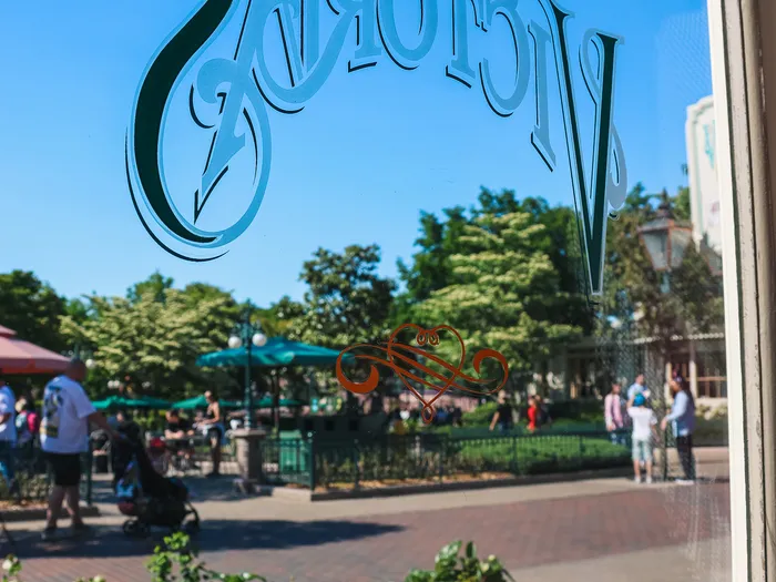 Vitrine gravée du nom Victoria’s reflétant la terrasse et les massifs fleuris de Main Street USA