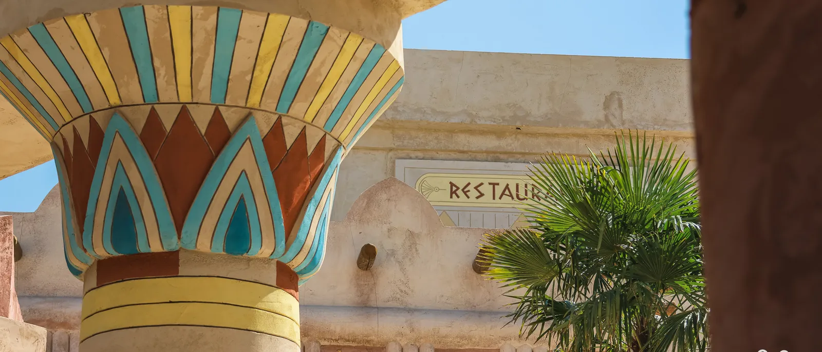 Chapiteau de colonne égyptienne peint en turquoise, jaune et brun, devant une façade beige portant l’enseigne Restaurant
