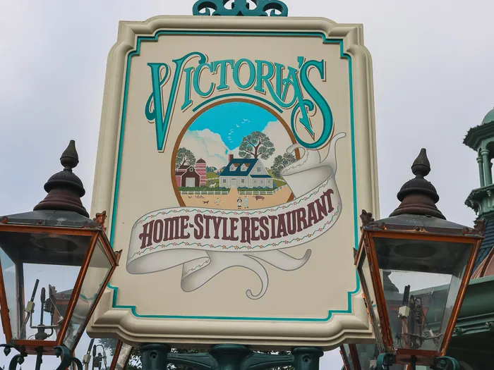 Gros plan sur l’enseigne peinte de Victoria’s Home-Style Restaurant, encadrée de deux lampadaires à Main Street USA.