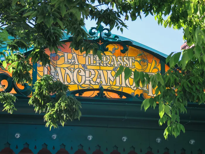 Orange Art Nouveau sign and green ironwork of La Terrasse Panoramique at Disneyland Paris, among the foliage