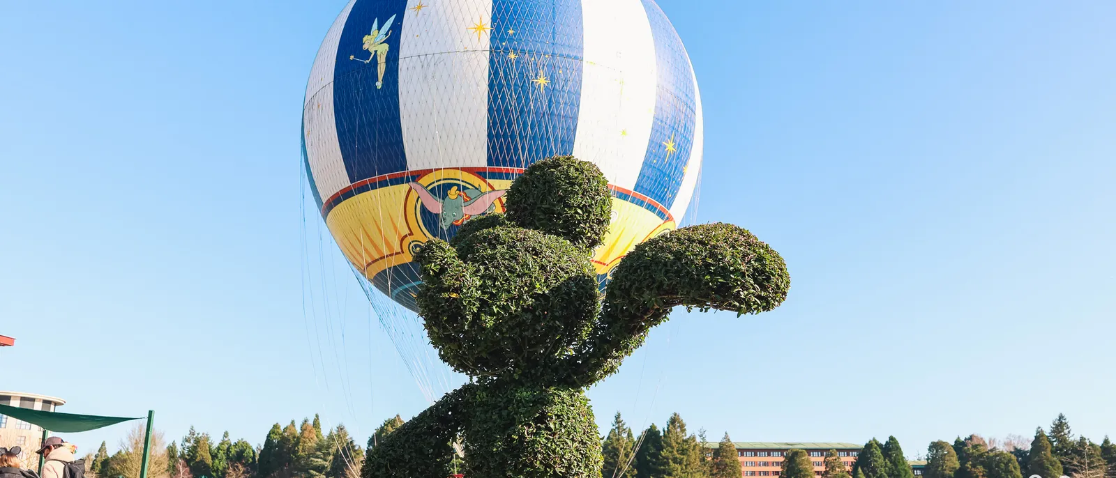Ballon captif blanc et bleu orné de la Fée Clochette, derrière un buisson taillé en forme de Mickey au Disney Village.
