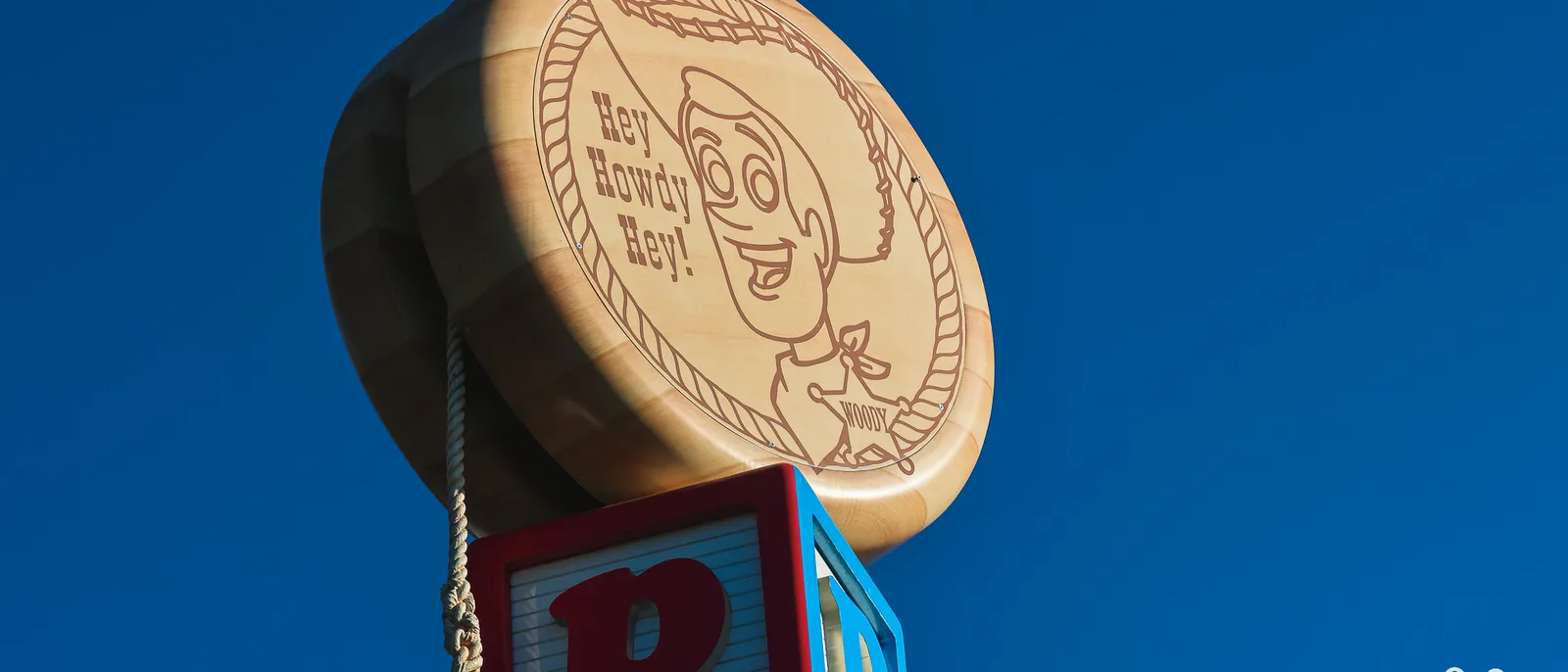 Yo-yo géant gravé « Hey Howdy Hey ! » à l’effigie de Woody, au-dessus d’un cube P, contre un ciel bleu, Toy Story Playland.