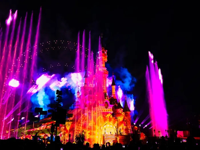Le château baigné de lumières orange et roses entre de hauts jets d’eau pendant Disney Tales of Magic au Parc Disneyland.