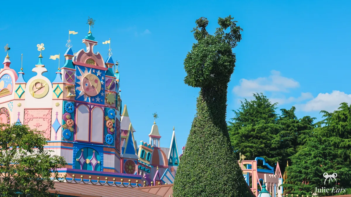The pastel facade of "it’s a small world" in Fantasyland, with its towers, flags and smiling clock face, behind a giraffe-shaped topiary, under a blue sky at Disneyland Paris