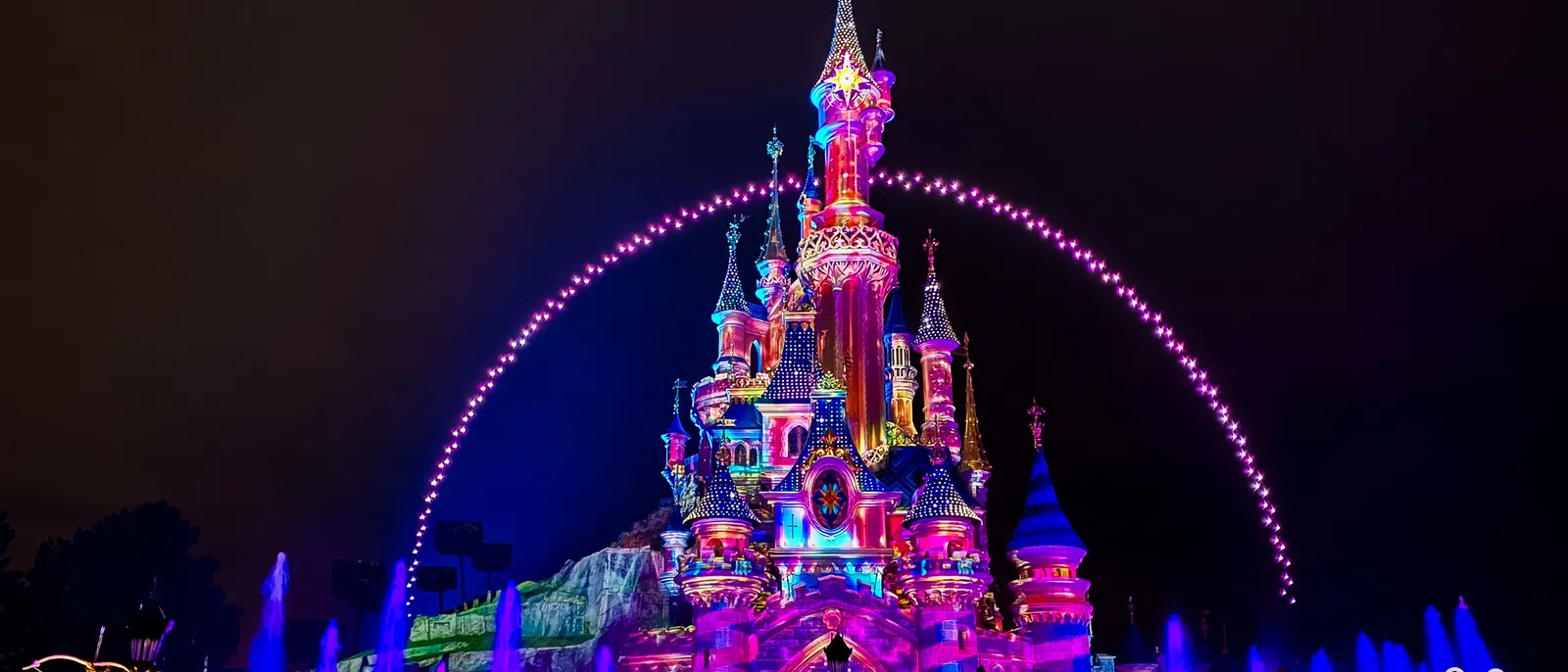 Arche de drones lumineux au-dessus du château illuminé, avec jets d’eau devant, la nuit à Disneyland Paris
