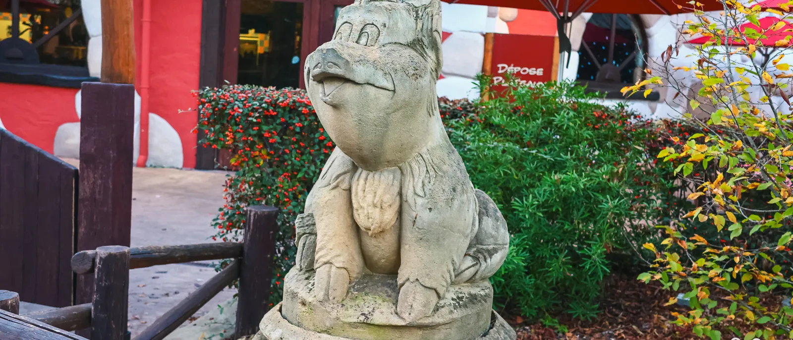 Statue en pierre d’un sanglier assis accoudé à un tonneau, devant une façade rouge et un parasol de terrasse