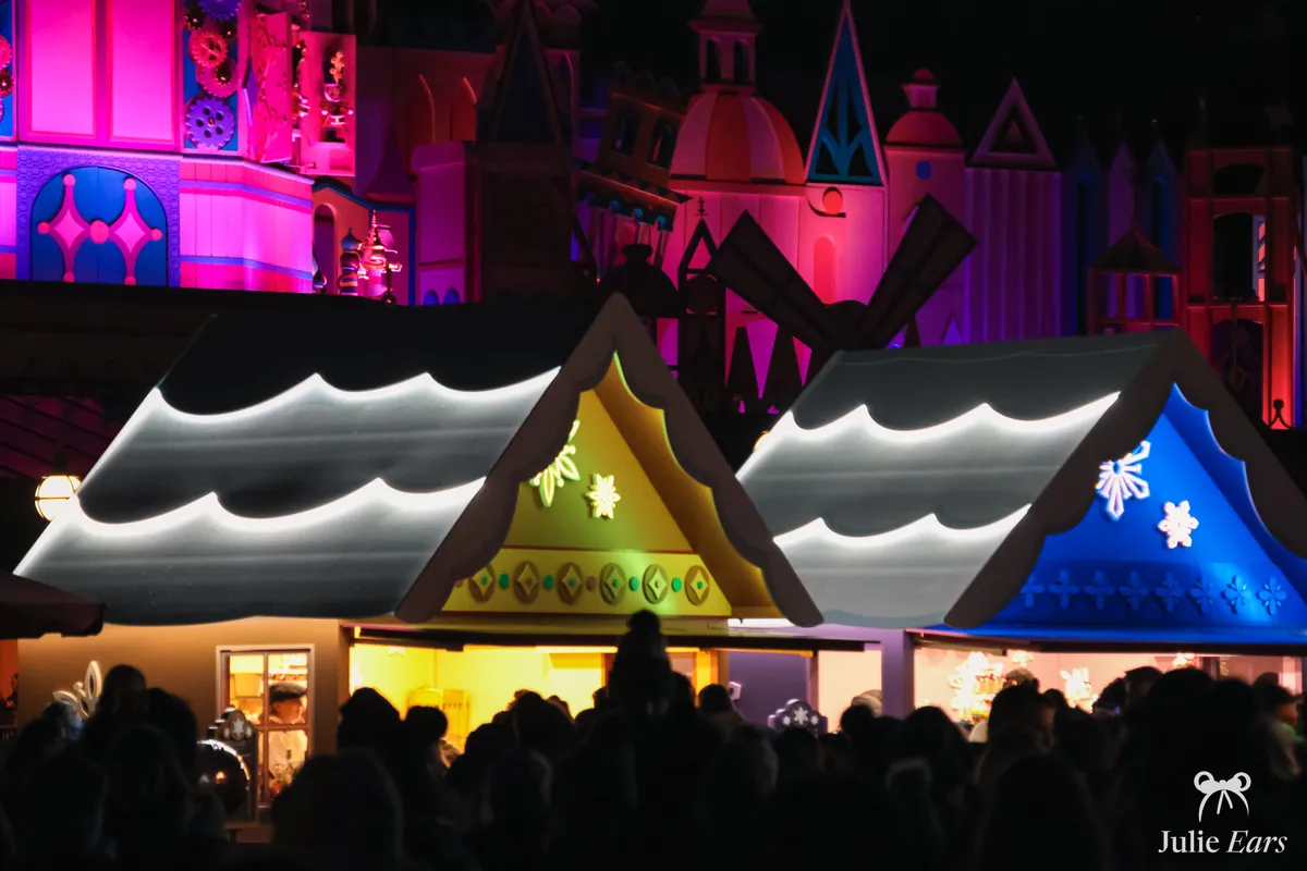 Illuminated festive food chalets of the L’Hiver Gourmand festival, in front of the "it’s a small world" frontage in Fantasyland, in the evening.
