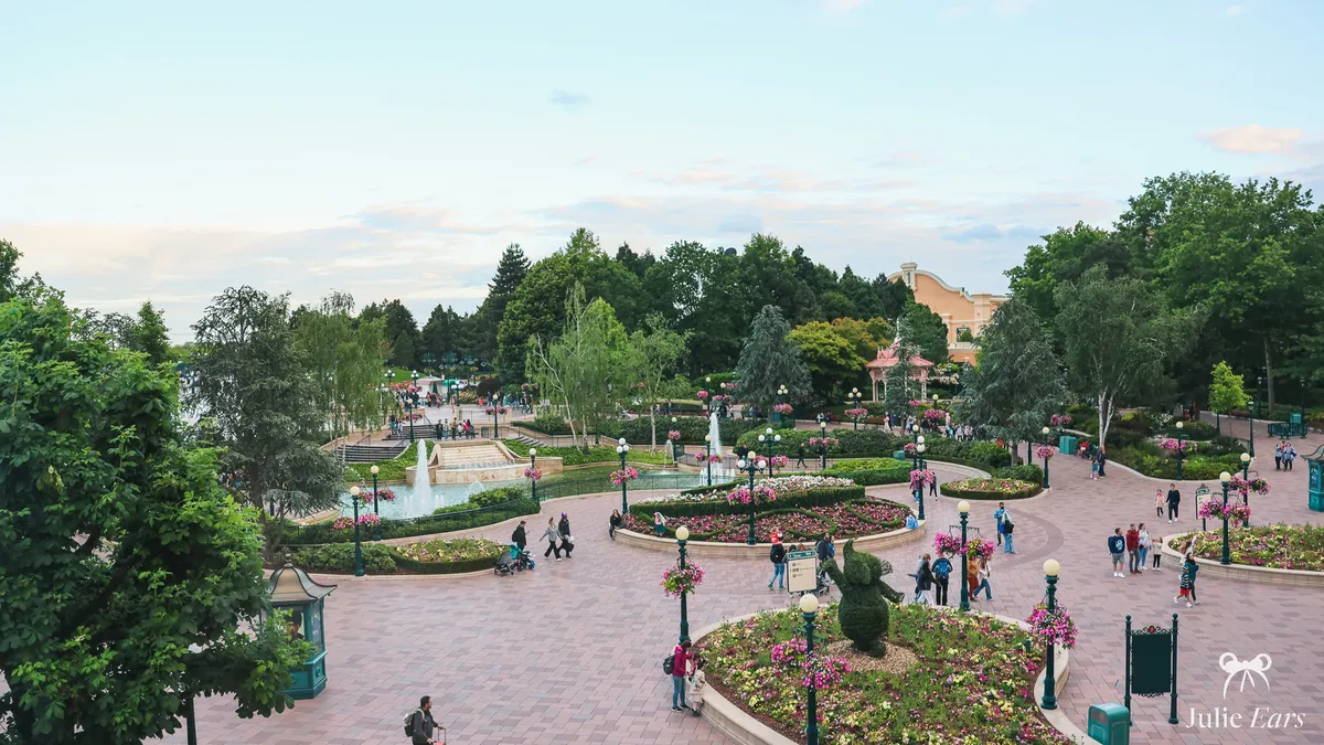 Disneyland Paris entrance esplanade seen from above at the end of the day, with paths, flower beds, fountain pools, a topiary and a few visitors