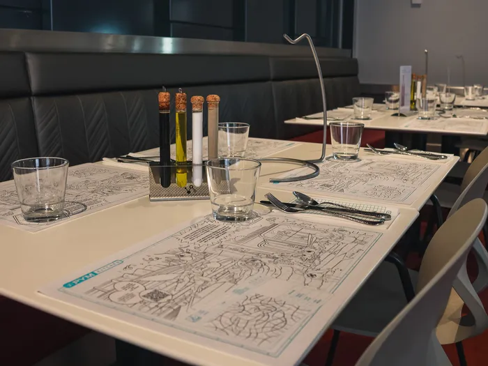 Tables du PYM Kitchen avec sets illustrés, banquette grise et chaises blanches sur sol rouge