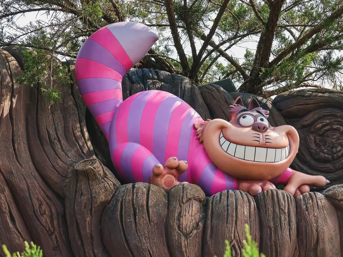 Statue du Chat du Cheshire rose et violet allongée sur un tronc rocheux à l’entrée du labyrinthe
