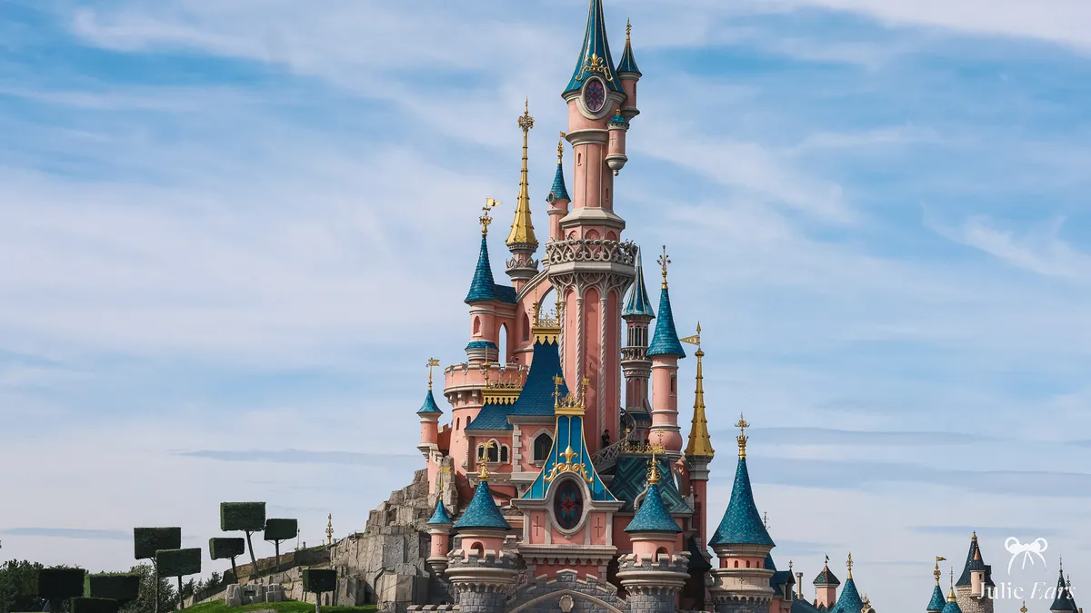 Sleeping Beauty Castle at Disneyland Paris, with its pink towers, blue roofs and golden spires, beside the rocky hill, under a hazy blue sky