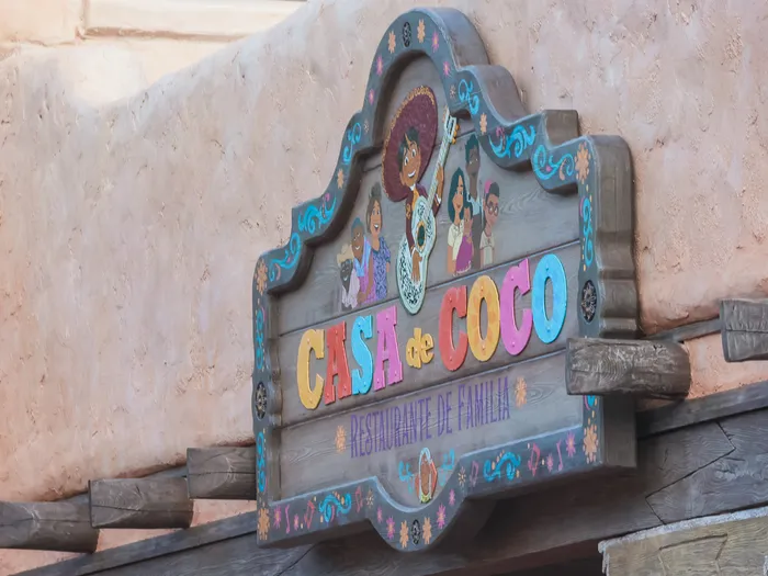 Enseigne en bois sculpté du restaurant Casa de Coco avec personnages de Coco, fixée sur un mur ocre à Frontierland