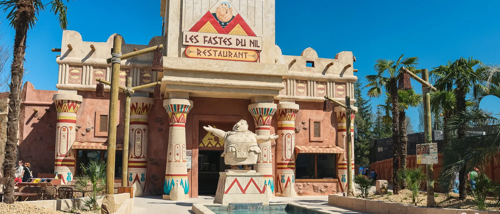 Façade ocre et rose du restaurant Les Fastes du Nil, colonnes peintes, statue égyptienne et bassin d’eau sous un ciel bleu