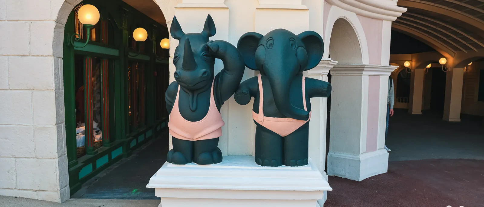 Statues d’un rhinocéros et d’un éléphant en maillot de bain rose sur un socle blanc, sous des arcades pastel