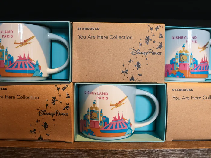 You Are Here Collection Disneyland Paris mugs from Starbucks, illustrated with the carousel and Space Mountain, lined up in their boxes on a shelf
