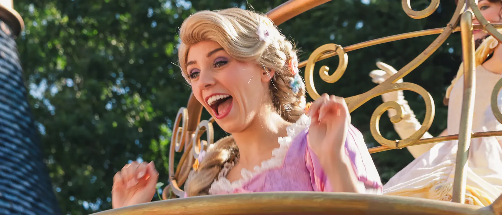 La princesse Raiponce, longue tresse blonde et robe mauve, salue en riant depuis un char de parade.