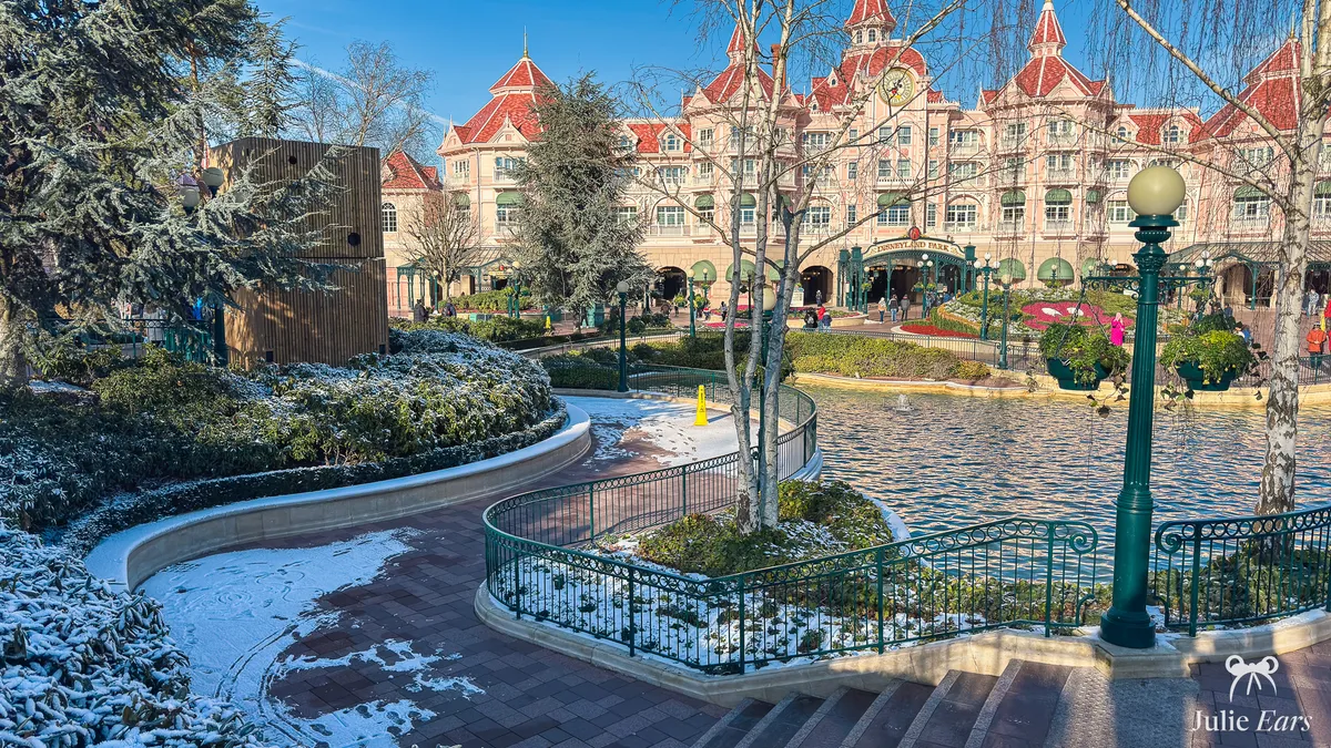 Snow-dusted gardens and paths by the Disneyland Hotel, with its ornamental pool, green lampposts and a yellow slippery surface sign, Disneyland Paris