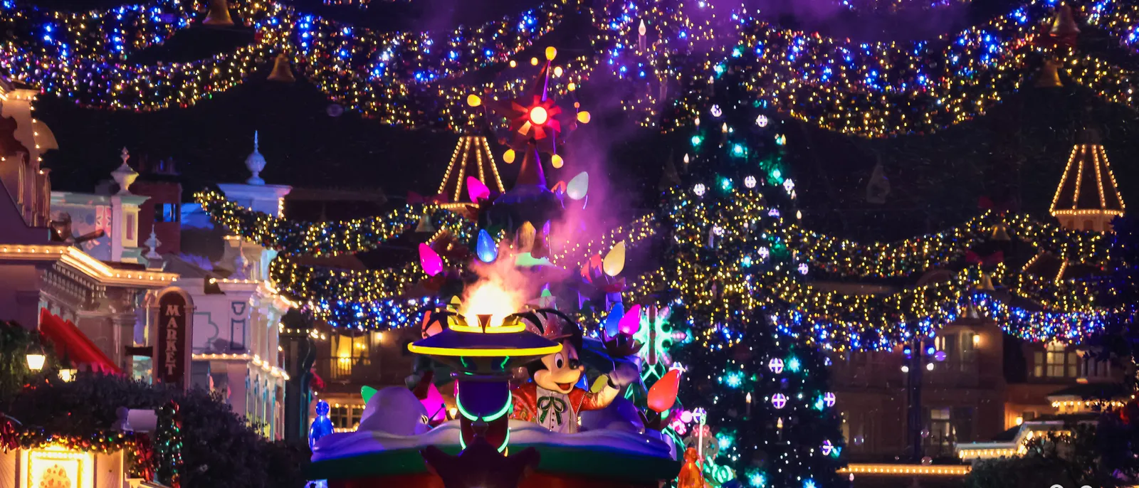 Char coloré avec Mickey défilant la nuit sous des guirlandes lumineuses bleues et dorées, sapin illuminé en fond.
