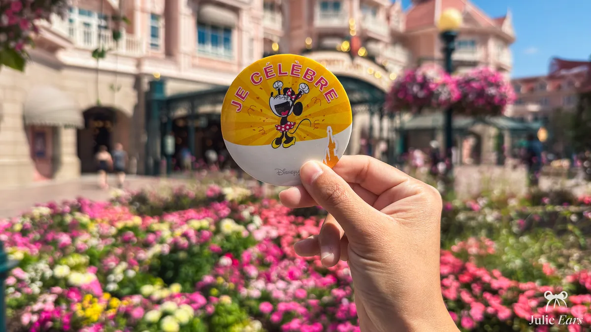 Main tenant le badge anniversaire « Je célèbre » de Disneyland Paris, avec Minnie, devant l’entrée du parc fleurie