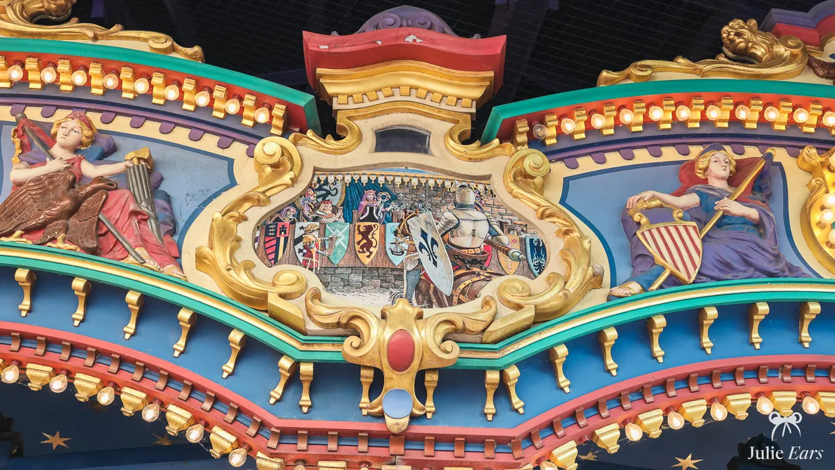 Painted, gilded pediment of Le Carrousel de Lancelot in Fantasyland: a jousting knight with a fleur-de-lis shield, coats of arms and strings of bulbs