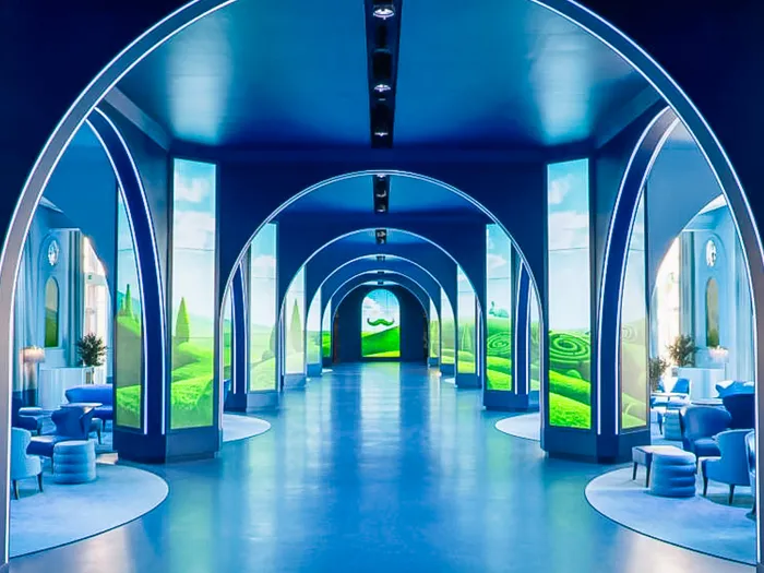 Galerie d’accueil aux arches bleues et écrans de collines vertes, avec fauteuils et topiaires, au Grand Magic Hotel.