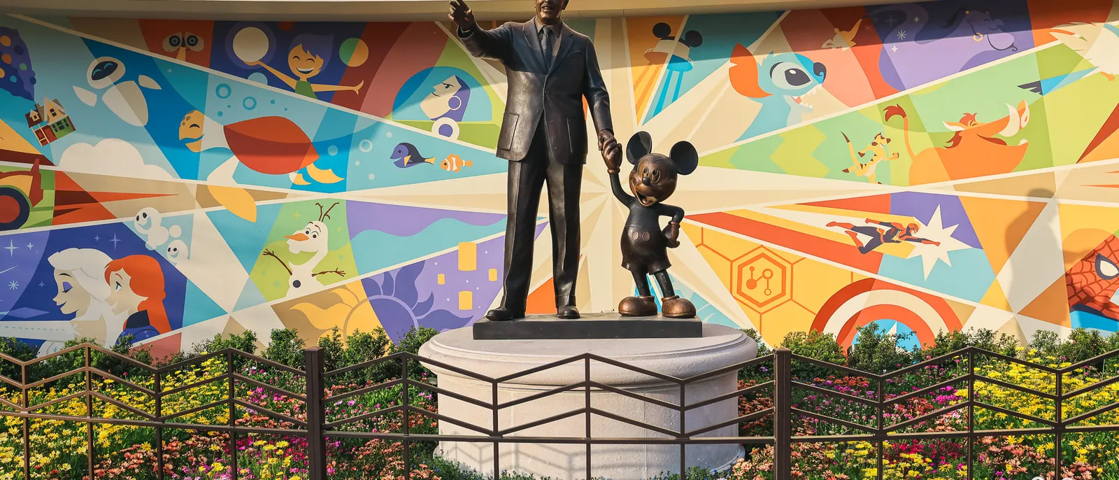 Statue en bronze de Walt Disney tenant la main de Mickey, devant une fresque colorée de personnages, à Disneyland Paris.