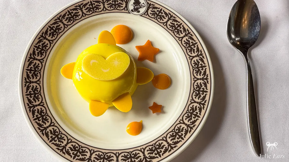 Un dessert en dôme jaune brillant, entouré de pétales, d’étoiles orange et de points de coulis, dans une assiette au logo Mickey du restaurant Walt’s, sur Main Street U.S.A. à Disneyland Paris