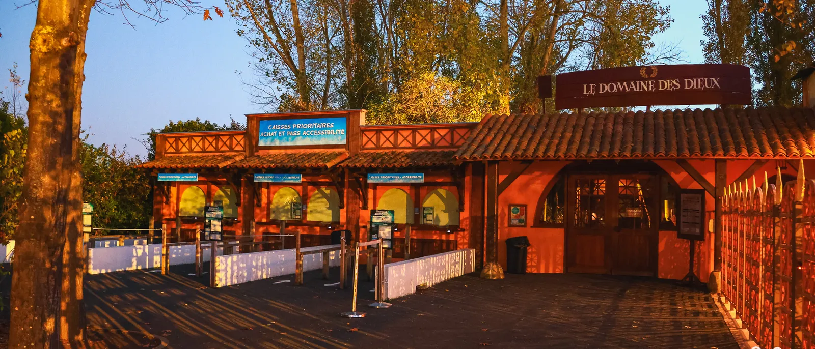 Entrée en bois rouge du Domaine des Dieux au soleil rasant, panneau bleu des caisses prioritaires devant les files vides