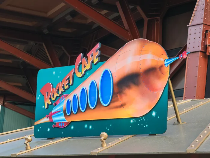 Enseigne peinte du Rocket Café avec une fusée orange filant sur fond étoilé, sous la charpente rouge de Discoveryland.