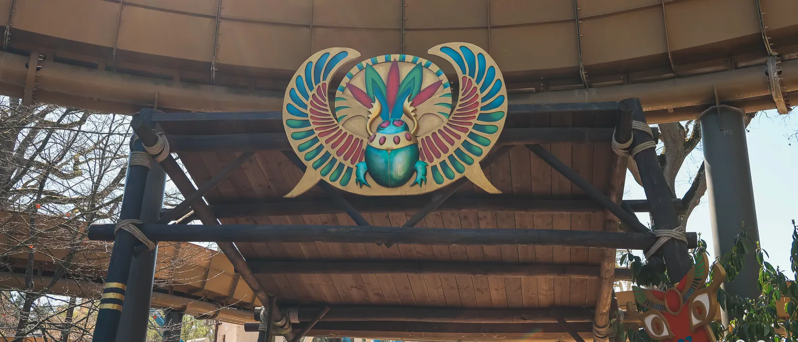 Scarabée ailé peint en bleu, rouge et or fixé sous une toiture en bois, décor égyptien du Parc Astérix