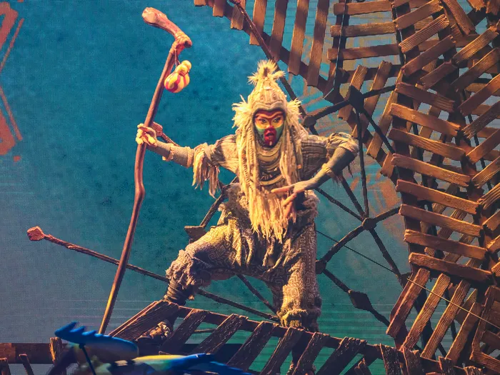 Comédien costumé en chaman du Roi Lion tenant un bâton sur une structure en bois, spectacle Mickey et le Magicien