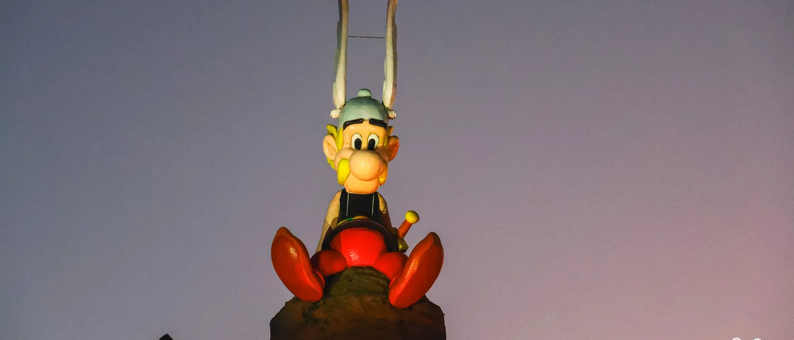 Statue d’Astérix assis au sommet d’un rocher, casque ailé et moustache jaune, se détachant sur un ciel violet de fin de journée
