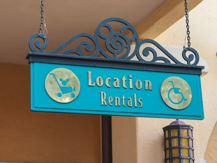 Turquoise wrought-iron hanging sign "Location / Rentals", decorated with gold pushchair and wheelchair medallions, at Disneyland Paris.